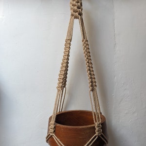 Puede incluir: Un colgador de plantas de macramé hecho a mano con un cuenco de madera. La cuerda beige está anudada para formar el colgador, que suspende un cuenco de madera marrón oscuro. El colgador está diseñado para colgarse de un gancho, añadiendo un toque bohemio.