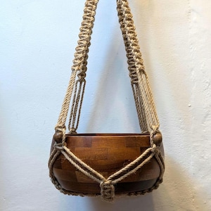 May include: A wooden hanging planter with a natural rope macrame design. The planter is made of dark wood and has a round shape.