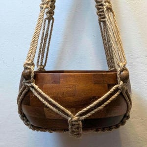 May include: A hanging wooden planter with a natural rope macrame design. The wooden bowl is made of various wood pieces and has a warm brown color. The rope is a light tan color and is knotted to create the hanging structure.