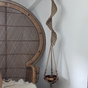 Pode incluir: Um suporte de plantas em macramé com um design em espiral, suspenso por um gancho de madeira escura. O suporte da planta é uma cesta tecida com uma borla. Uma cadeira de pavão de vime está no fundo.