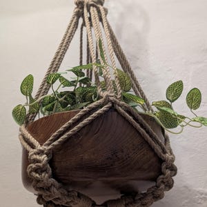 Pode incluir: Um suporte de plantas suspenso em macramé com uma tigela de madeira contendo uma planta verde com veias brancas. O macramé é feito de corda bege e é amarrado no topo e ao redor da tigela. O suporte de plantas está suspenso contra uma parede branca.