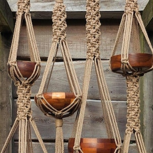 May include: Four macrame plant hangers with wooden bowls. The hangers are made of tan rope and feature intricate knotting patterns. The wooden bowls are a rich brown color and appear to be empty. The background is a weathered wooden wall.