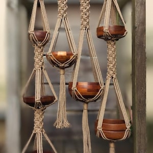 May include: A selection of macrame plant hangers, each crafted with beige rope. The hangers cradle wooden bowl planters, displaying natural wood grains. The planters are of varying sizes and shapes. The background is a blurred wooden structure.
