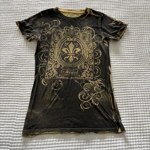 May include: A black short-sleeve t-shirt featuring a gold-colored graphic print of a fleur-de-lis inside a rectangular ornate frame. The word Sinful is printed in a script font below the emblem, surrounded by swirling filigree patterns.