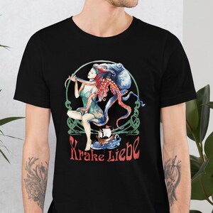 May include: Black t-shirt featuring a colorful illustration of a woman with red hair and octopus tentacles, holding a bow and arrow. The design includes a ship and the text "Krake Liebe" in red. The artwork is set within a green and white circular frame.