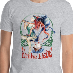 May include: A light gray t-shirt featuring a colorful illustration. The graphic depicts a woman with an octopus, a ship, and the text "Krake Liebe" in red. The design is framed by a green Celtic knot.