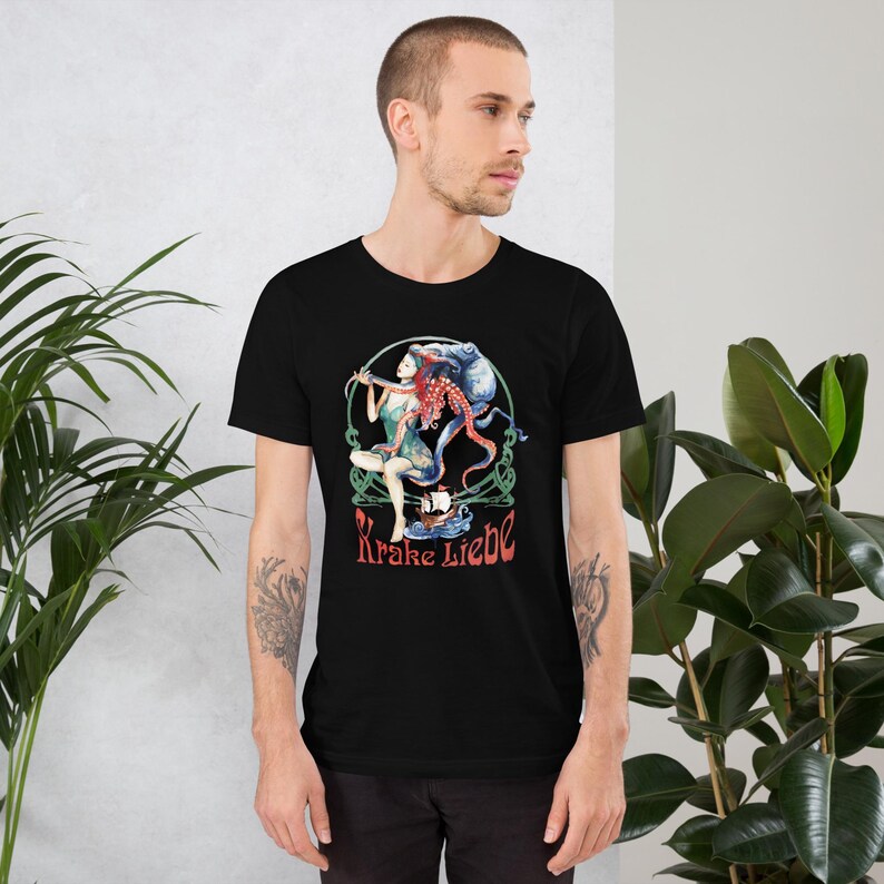 May include: Black t-shirt featuring a colorful illustration of a woman with red hair and octopus tentacles, holding a bow and arrow. The design includes a ship and the text "Krake Liebe" in red. The artwork is set within a green and white circular frame.