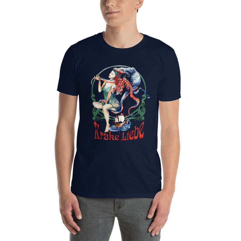 May include: Navy blue t-shirt featuring a colorful illustration. The graphic includes a woman, an octopus, and a ship, with the text "Krake Liebe" in red. The design is set within a green Celtic-style border.