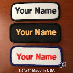 May include: Three embroidered name tags with the text "Your Name" in different colors and styles. The top tag is white with black text, the middle tag is black with gold text, and the bottom tag is white with red text. All tags have a blue border and are 1.5 inches by 4 inches. Made in the USA.