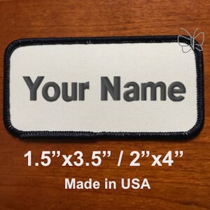 May include: White rectangular patch with black stitching and a black border. The patch has the text "Your Name" in gray embroidery. The patch is 1.5 inches by 3.5 inches or 2 inches by 4 inches. The text "Made in USA" is printed below the patch.