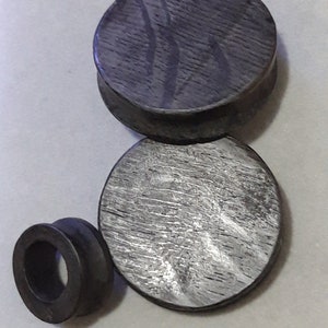 May include: Three wooden ear gauges in different sizes. The largest gauge is dark grey with a wood grain pattern. The medium gauge is a lighter grey with a wood grain pattern. The smallest gauge is black and round.