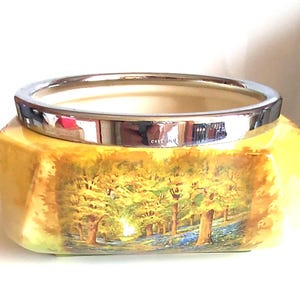 Puede incluir: Un cuenco de cerámica amarillo con un borde plateado y una escena pintada de un bosque con árboles y flores azules.