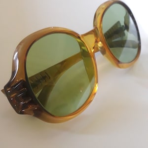 May include: Vintage oversized sunglasses with a light brown frame and green lenses. The sunglasses have a unique, rounded shape and a thick frame. The lenses are a light green color, and the frame has a slight gradient.