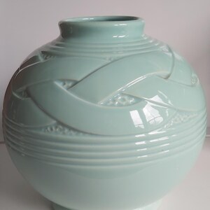 May include: A pale blue ceramic vase with a rounded form and a slender neck. The vase has a woven design around its midsection and horizontal lines near the base. The vase is approximately 25 cm tall.