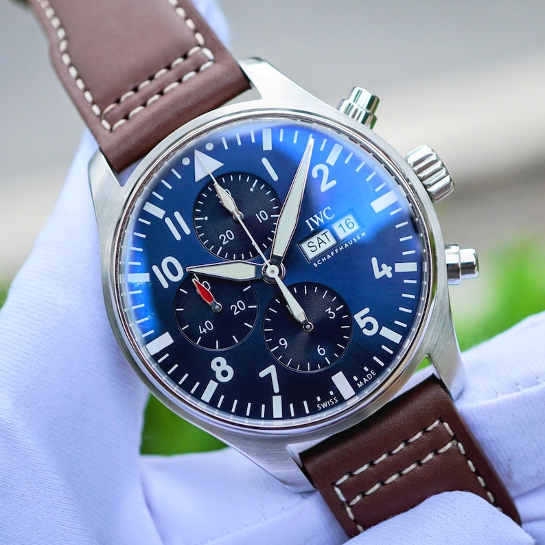 May include: A stainless steel IWC Schaffhausen chronograph watch with a deep blue dial, white Arabic numerals, and three sub-dials. It features a day-date window at the three o'clock position and a brown leather strap with white contrast stitching.