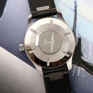 May include: A stainless steel IWC Pilot's watch with a black leather strap. The case back features an engraving of an airplane, the text International Watch Co. Pilot's Watch, and a 10 bar water resistance rating. The IWC logo is on the strap.