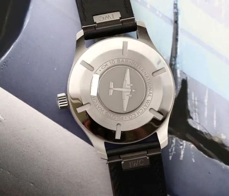 May include: A stainless steel IWC Pilot's watch with a black leather strap. The case back features an engraving of an airplane, the text International Watch Co. Pilot's Watch, and a 10 bar water resistance rating. The IWC logo is on the strap.
