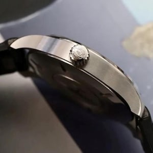 May include: A silver-toned wristwatch with a black strap, featuring a circular metallic case and a side-mounted winding crown with a textured edge. The watch case has a polished finish, and the crown displays a small engraved logo on its side.