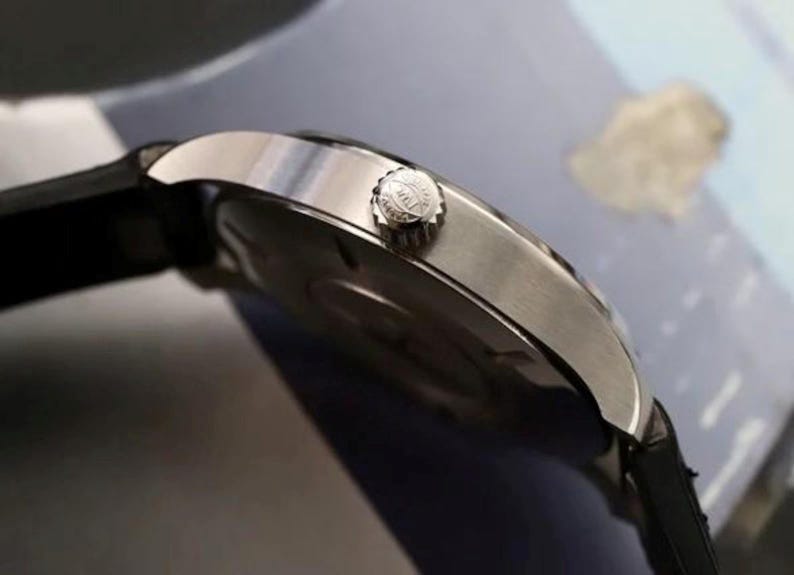 May include: A silver-toned wristwatch with a black strap, featuring a circular metallic case and a side-mounted winding crown with a textured edge. The watch case has a polished finish, and the crown displays a small engraved logo on its side.