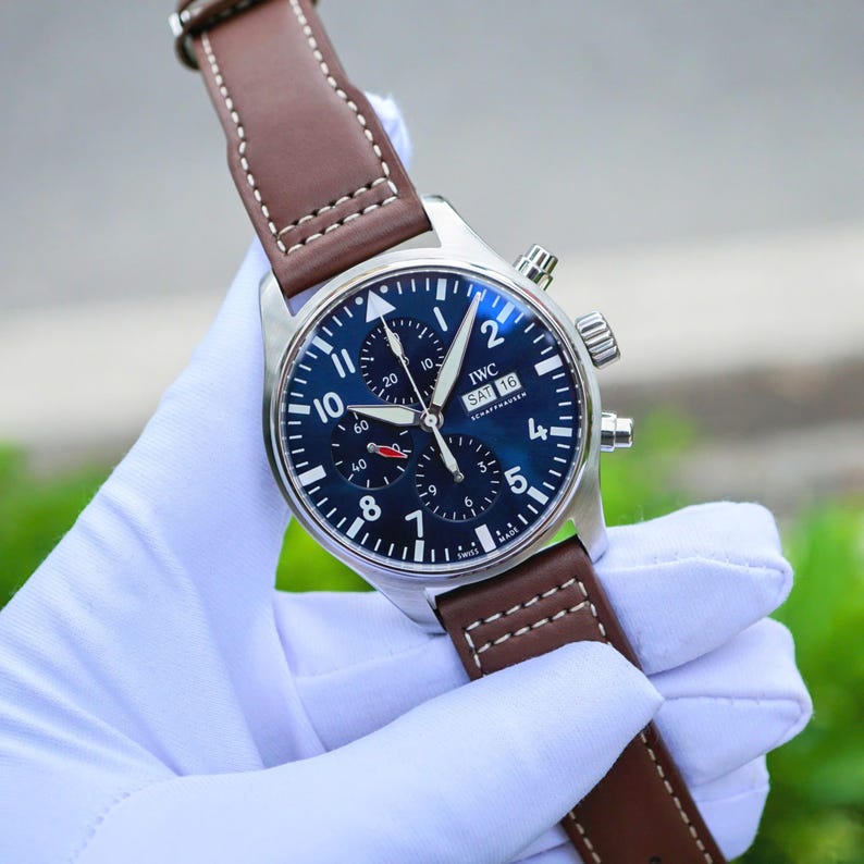 May include: A stainless steel IWC Schaffhausen chronograph watch with a dark blue dial, white Arabic numerals, and three sub-dials. The watch features a day-date window at the three o'clock position and a brown leather strap with white stitching.