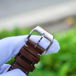 May include: A brown leather watch strap featuring a silver-toned metal buckle with the engraved text IWC. The strap includes two leather keepers and white contrast stitching along the edges, showcasing a classic design for a timepiece.