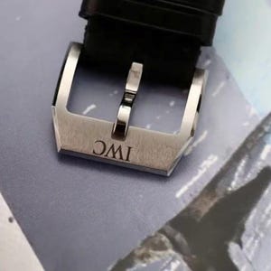 May include: A silver-toned stainless steel watch buckle featuring a brushed finish and the engraved letters IWC. The buckle is attached to a black textured leather watch strap, showcasing a classic tang style design with a polished metal pin.