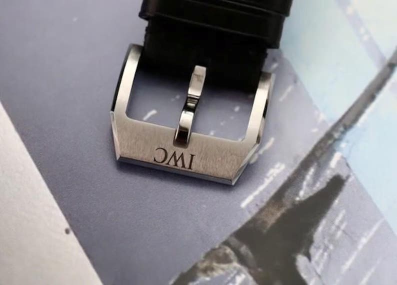 May include: A silver-toned stainless steel watch buckle featuring a brushed finish and the engraved letters IWC. The buckle is attached to a black textured leather watch strap, showcasing a classic tang style design with a polished metal pin.