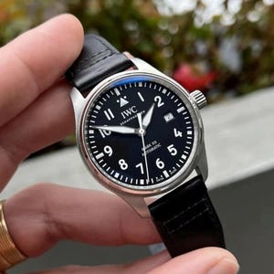 May include: An IWC Schaffhausen Mark XX automatic wristwatch with a round silver-tone stainless steel case, a black dial featuring white Arabic numerals, a date window at the three o'clock position, and a black leather strap with stitching.
