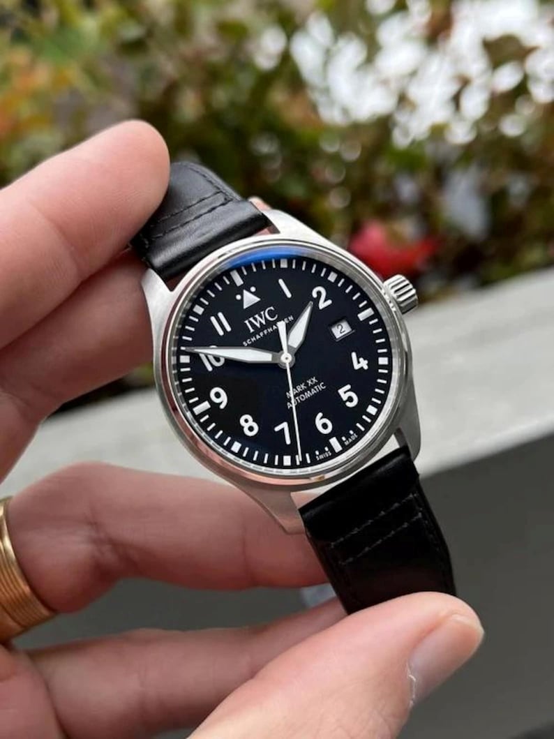 May include: An IWC Schaffhausen Mark XX automatic wristwatch with a round silver-tone stainless steel case, a black dial featuring white Arabic numerals, a date window at the three o'clock position, and a black leather strap with stitching.
