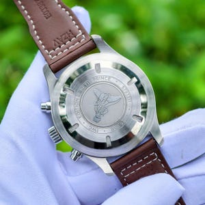 May include: A stainless steel watch case back with a brown leather strap. The case back features an engraving of a character, the text EDITION LE PETIT PRINCE, IWC, 6 BAR, and a serial number. The strap has white stitching and visible text.