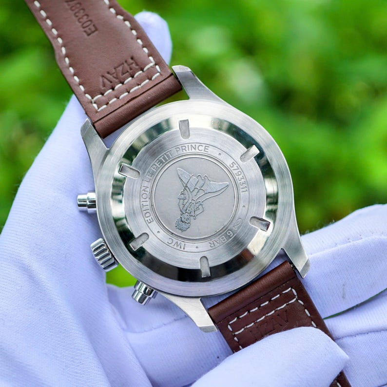 May include: A stainless steel watch case back with a brown leather strap. The case back features an engraving of a character, the text EDITION LE PETIT PRINCE, IWC, 6 BAR, and a serial number. The strap has white stitching and visible text.