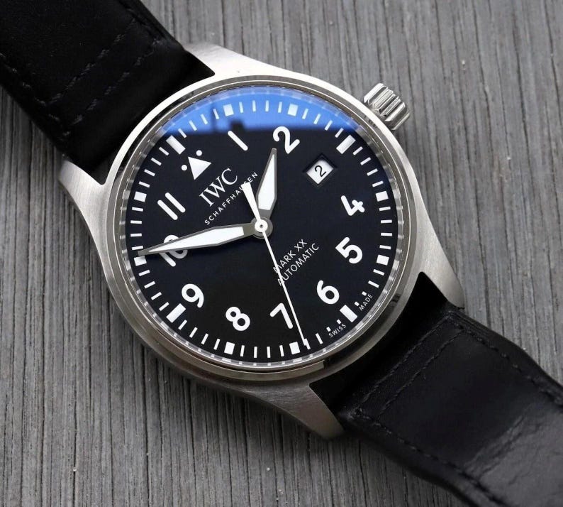 May include: An IWC Schaffhausen Mark XX Automatic wristwatch with a round stainless steel case, a black dial featuring white Arabic numerals, luminous hands, a date window at the three o'clock position, and a black leather strap.