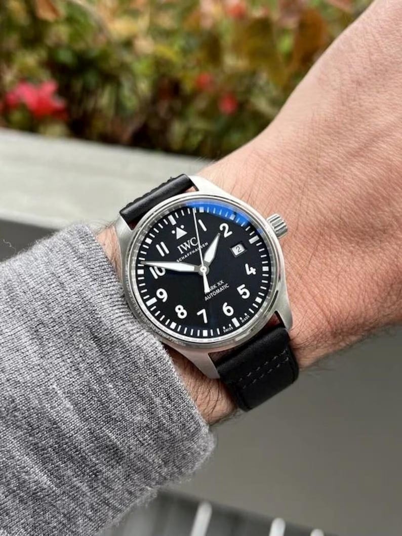 May include: An IWC Mark XX automatic wristwatch with a round stainless steel case, a black dial featuring white Arabic numerals, a date window at the three o'clock position, and a black leather strap with visible stitching.
