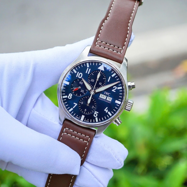 May include: A stainless steel chronograph wristwatch with a deep blue dial, white Arabic numerals, and three sub-dials. The watch features a day-date display at the three o'clock position, luminous hands, and a brown leather strap with white stitching.