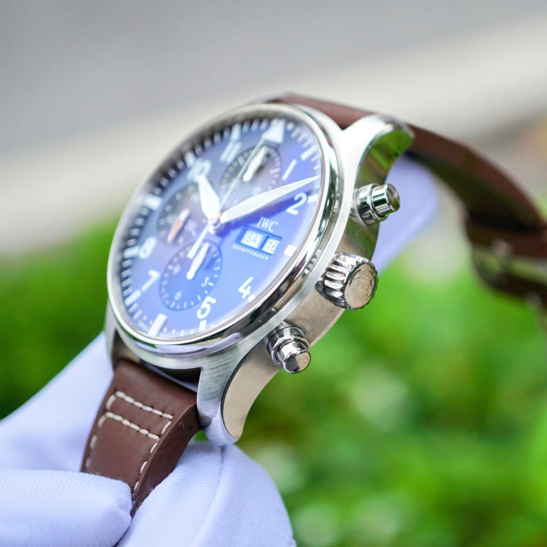 May include: A silver-tone chronograph wristwatch with a blue dial, white Arabic numerals, and a brown leather strap featuring white contrast stitching. The watch face displays the IWC brand name, a day-date window, and three sub-dials.