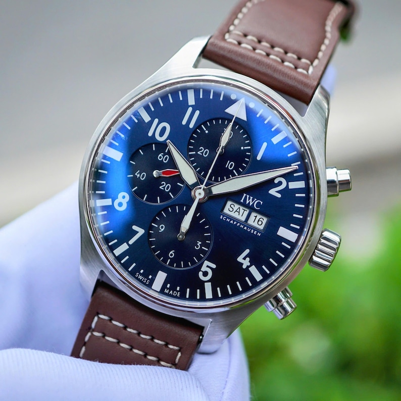 May include: A stainless steel IWC Schaffhausen chronograph watch with a deep blue dial, white Arabic numerals, three sub-dials, and a day-date window. The watch features luminous hands and is attached to a brown leather strap with white stitching.