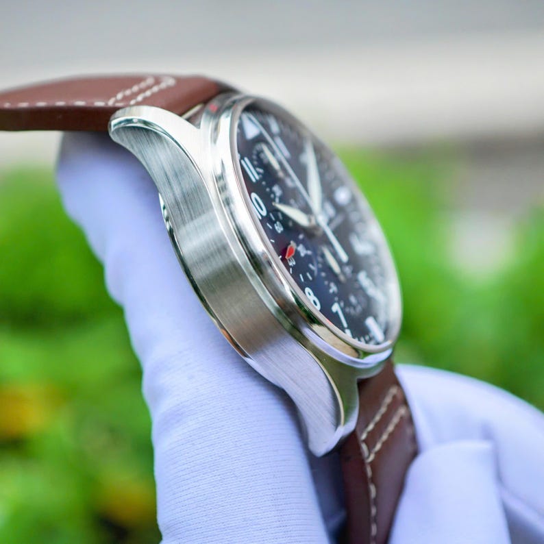 May include: A wristwatch with a brushed silver-tone metal case and a dark brown leather strap featuring white stitching. The watch face is black with white Arabic numerals, silver hands, and a small red accent on the dial markers.