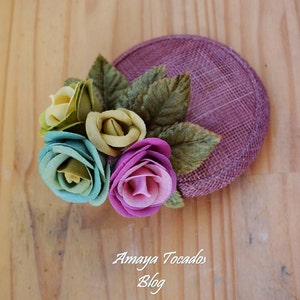 Könnte beinhalten: Ein dekoratives Haarteil mit einem kreisförmigen, mauvefarbenen Grund, verziert mit Stoffrosen in Grün, Blau und Rosa sowie grünen Stoffblättern. Der Text "Amaya Tocados Blog" ist auf der Holzoberfläche sichtbar.