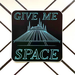 May include: Black and white sticker with a blue outline. The sticker features a stylized outline of the Space Mountain ride at Disney World and the text "GIVE ME SPACE".