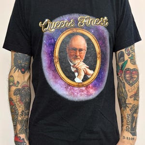 May include: Black t-shirt featuring a portrait in a gold frame with the text "Queens Finest". The portrait is set against a purple and blue galaxy background. The t-shirt is a crew neck style.