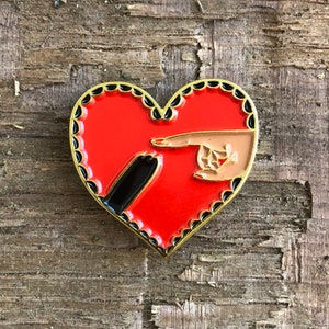 Op de afbeelding: Rode hartvormige emaille pin met een zwart-witte hand die naar rechts wijst en een bruine hand die naar links wijst. De pin heeft een gouden rand en zwarte gegolfde randen.