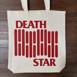May include: Cream-coloured canvas tote bag featuring the words "DEATH STAR" in red, bold, sans-serif font. Below "DEATH" is a series of vertical red lines. The bag has two matching canvas handles. The design is printed on the front.