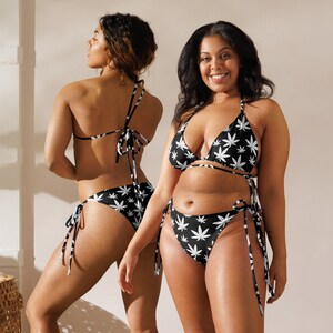 May include: Two women wearing black and white bikini tops and bottoms with a marijuana leaf print. The bikini tops are triangle style and the bottoms are tie-side.