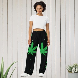 Puede incluir: Pantalón negro con un estampado de hojas de marihuana verdes. El pantalón tiene una cintura elástica y un corte holgado.