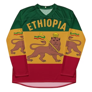 Puede incluir: Una camiseta de manga larga con un diseño verde, amarillo y rojo. La camiseta presenta la bandera de Etiopía con un león y la palabra "ETHIOPIA" impresa en la parte delantera.