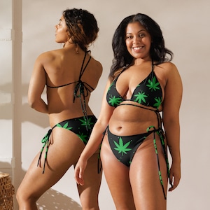 May include: Two women wearing black and green bikini tops and bottoms with a marijuana leaf print. The bikini tops are halter style and the bottoms are high-cut.