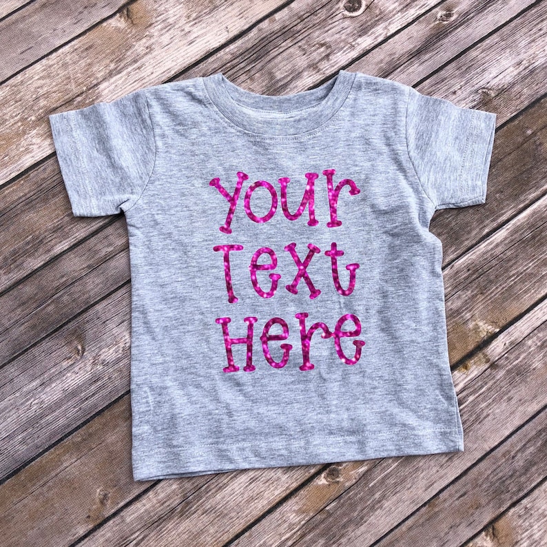 May include: A gray t-shirt with the text "YOUR TEXT HERE" in pink glitter lettering.