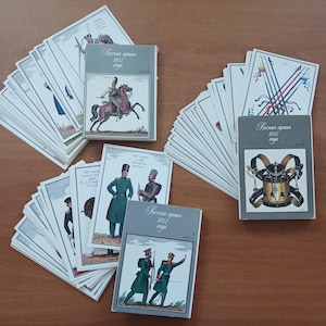 Może przedstawiać: Trzy stosy ilustrowanych kart zatytułowanych "Русская армия 1812 года", przedstawiających historyczne postacie wojskowe, sprzęt i broń. Każdy stos jest zwieńczony kartą przedstawiającą żołnierzy na koniach, bęben z paskami lub żołnierzy w mundurach.