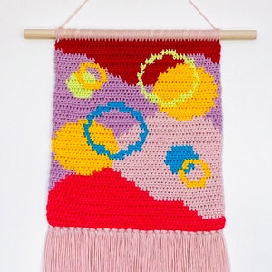 Könnte beinhalten: Ein farbenfroher gehäkelter Wandbehang mit einem rosa Fransenbesatz. Der Wandbehang zeigt abstrakte Formen in Rot, Gelb, Lila und Blau.
