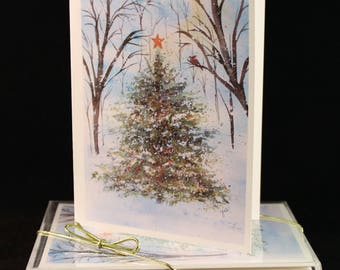 May include: A set of Christmas cards featuring a watercolor painting of a snowy forest scene with a pine tree, a red star, and a cardinal perched on a branch. The cards are packaged in a clear plastic box with a gold ribbon.